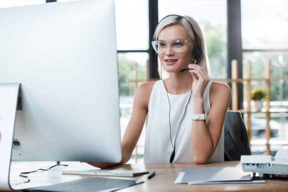 selective focus of cheerful blonde woman in headset working in office near computer monitor